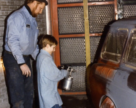 A man and young boy painting the rear panel of an old car in a garage