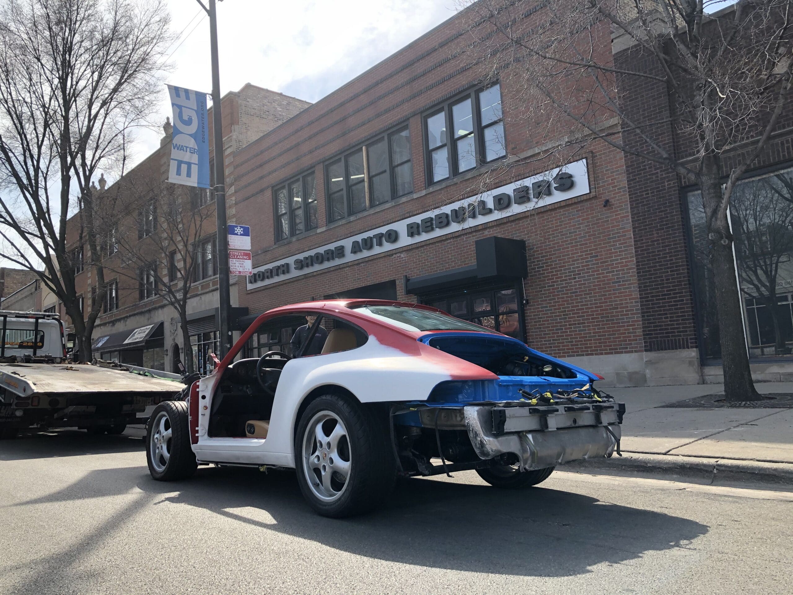 A broken-down car with a damaged body parked on the roadside, surrounded by grass and trees.