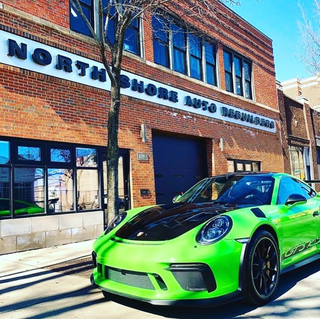 A green Porsche parked in front of a red brick building, showcasing its sleek design against the textured backdrop.